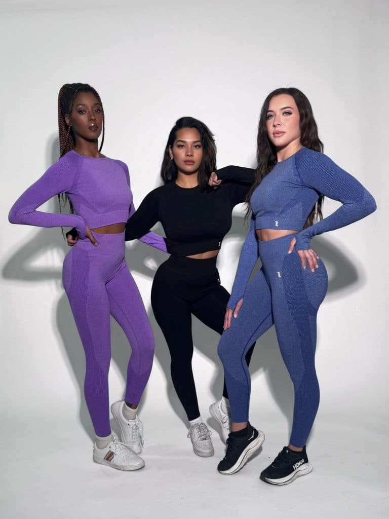 Three women pose confidently in Allure Set V2 activewear—matching long-sleeve crop tops and high-waisted leggings in purple, black, and blue—standing against a plain white background.