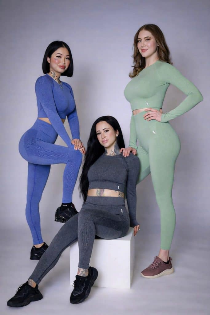 Three women wear the Allure Set V2 activewear in blue, gray, and green while posing in a studio—one standing left in blue, one sitting center in gray on a white block, and one standing right in green—all with sneakers, facing the camera.