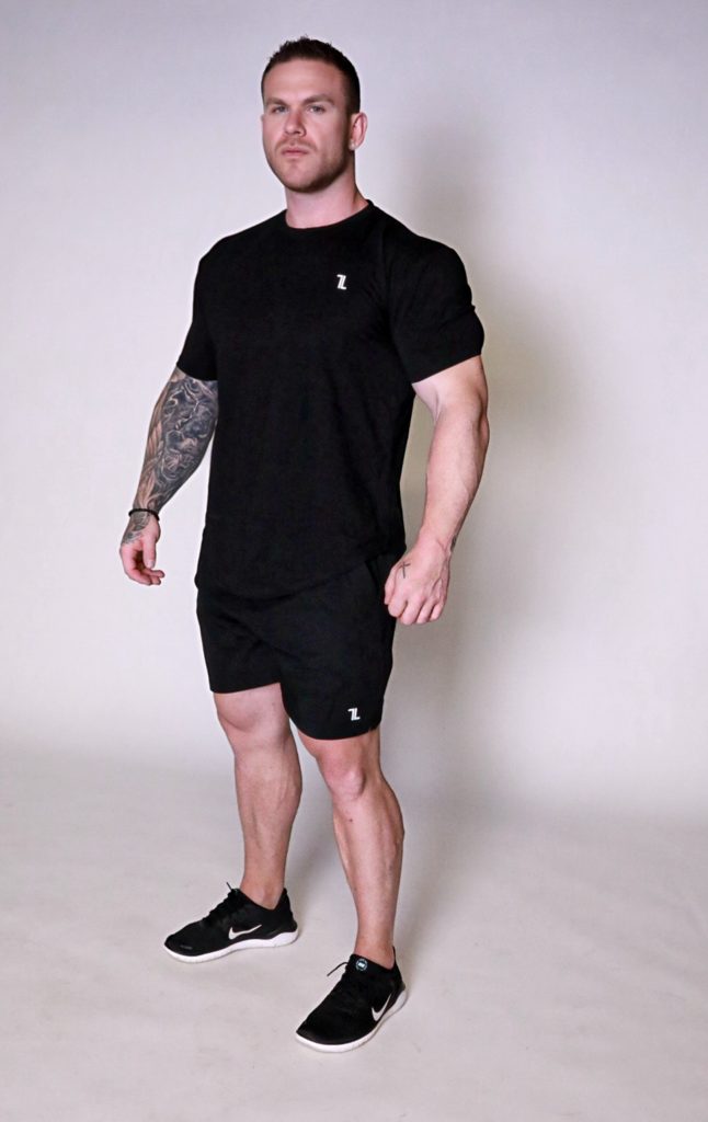 A muscular man with short hair and a tattoo sleeve on his left arm stands confidently in a studio, wearing the Genesis Tee V2, black shorts, and black Nike sneakers, looking slightly upwards.