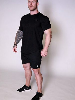A muscular man with short hair and a tattoo sleeve on his left arm stands confidently in a studio, wearing the Genesis Tee V2, black shorts, and black Nike sneakers, looking slightly upwards.