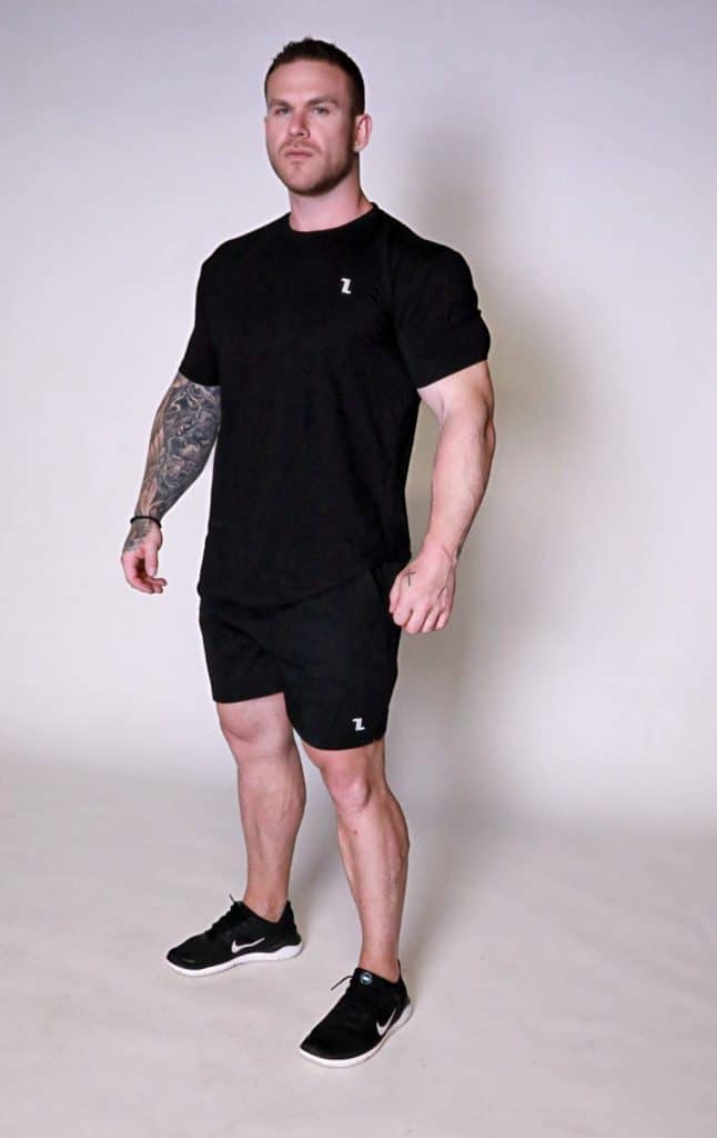 A muscular man with short hair and a tattoo sleeve on his left arm stands confidently in a studio, wearing the Genesis Tee V2, black shorts, and black Nike sneakers, looking slightly upwards.