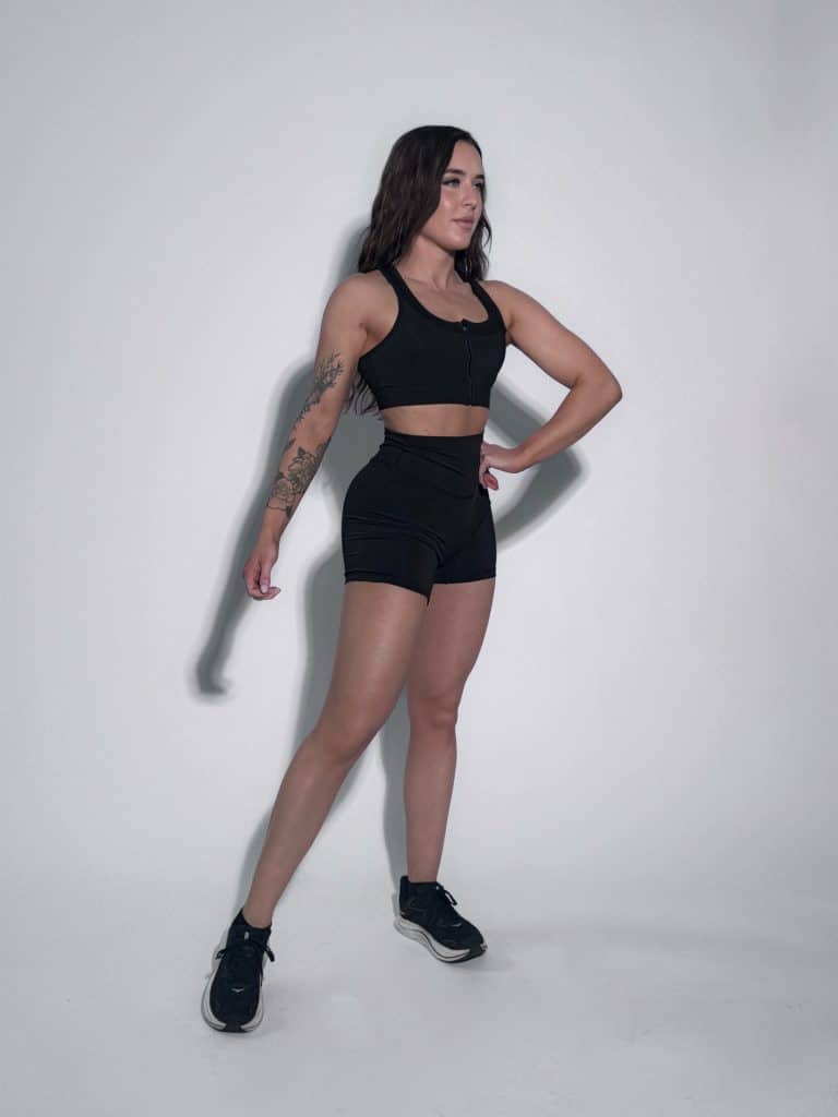 A woman in black athletic wear and sneakers stands against a plain white background, posing with one hand on her hip and the other relaxed by her side. She has visible tattoos on her left arm.