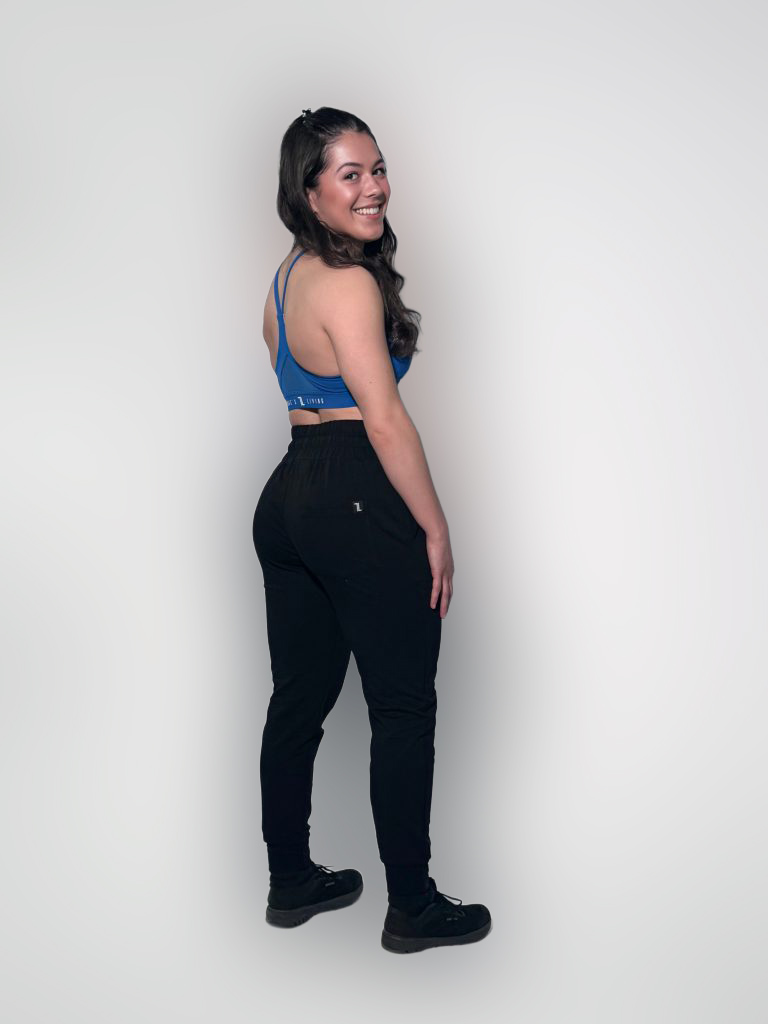 A woman with long dark hair, wearing a blue sports bra, black pants, and black shoes, stands and smiles at the camera against a plain light background. She is turned slightly to the side with her hands by her hips.