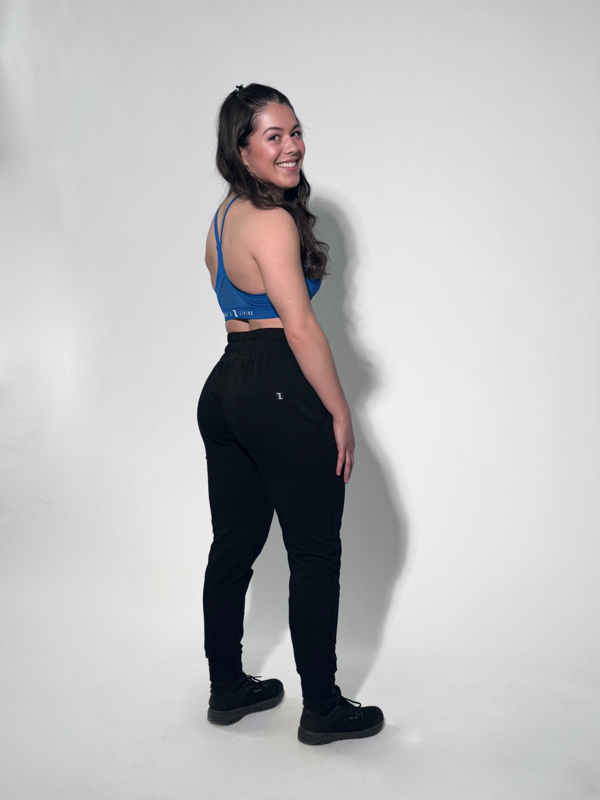Black 1 A woman with long dark hair, wearing a blue sports bra, black pants, and black shoes, stands and smiles at the camera against a plain light background. She is turned slightly to the side with her hands by her hips.