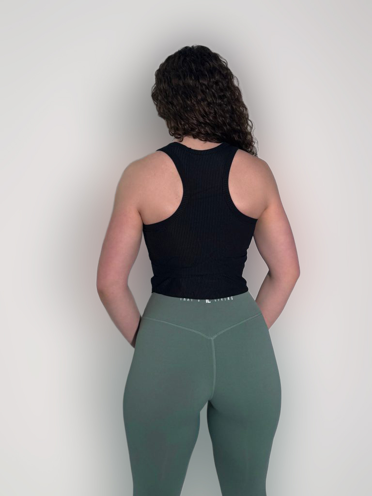 A person with curly hair faces away from the camera, wearing the LuxeForm Tank in black and green leggings, hands resting behind their back against a plain backdrop.