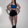 A woman stands with her hands on her hips, wearing GLYDE Shorts, a blue sports bra, and black athletic shoes. She looks to the side against a plain white background.