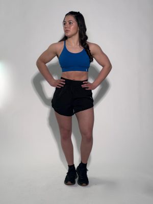 A woman stands with her hands on her hips, wearing GLYDE Shorts, a blue sports bra, and black athletic shoes. She looks to the side against a plain white background.