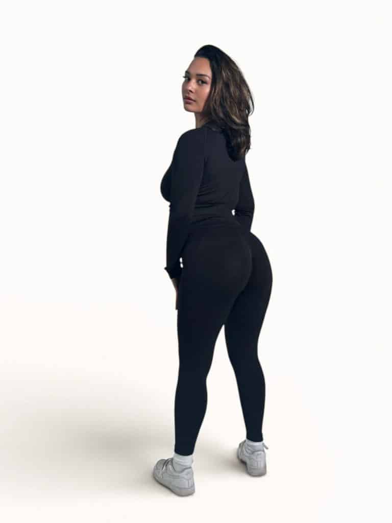 A woman with medium-length brown hair, wearing a black long-sleeve top, black leggings, and white sneakers, stands on a plain white background, looking over her shoulder at the camera.