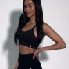 A woman with long dark hair stands against a plain white background, wearing a black sports bra and high-waisted leggings, smiling slightly with one hand on her hip.