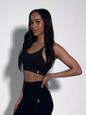 A woman with long dark hair stands against a plain white background, wearing a black sports bra and high-waisted leggings, smiling slightly with one hand on her hip.