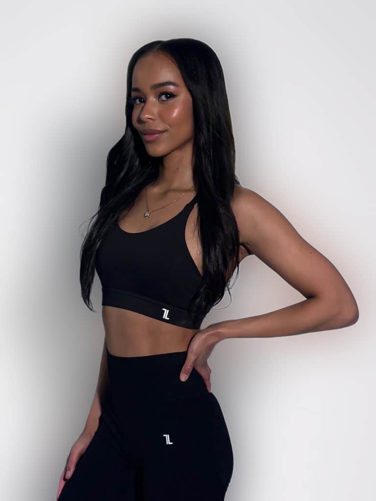 A woman with long dark hair stands against a plain white background, wearing a black sports bra and high-waisted leggings, smiling slightly with one hand on her hip.