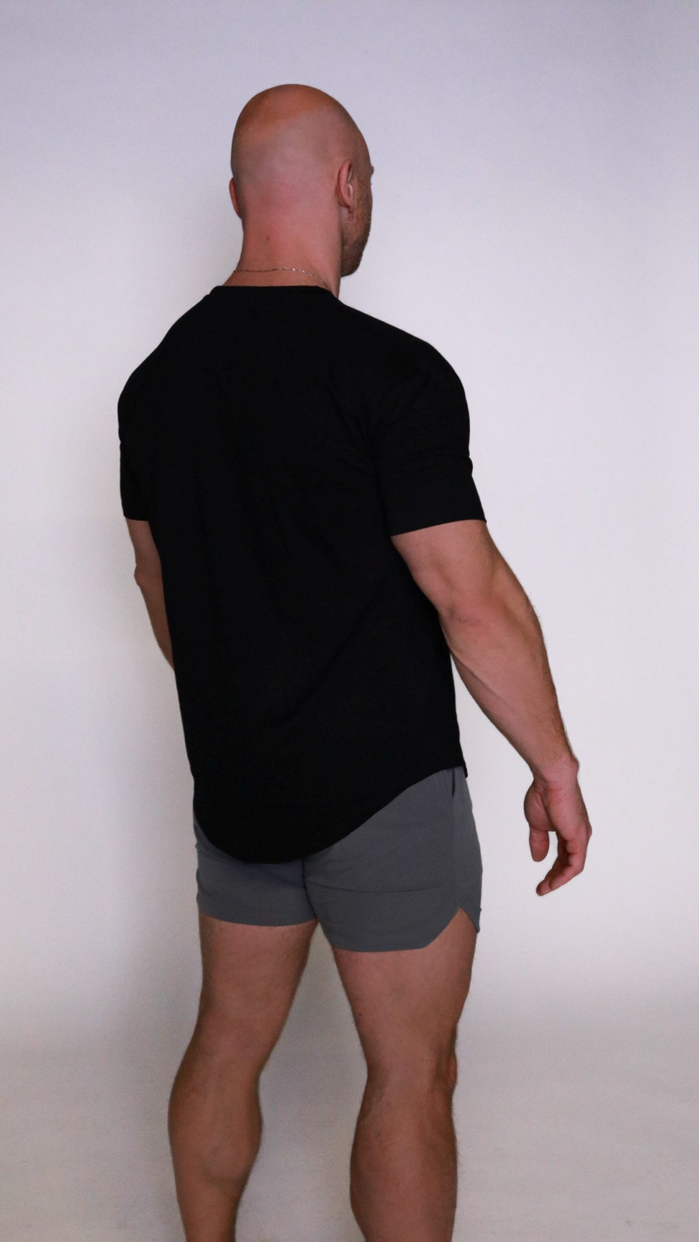 A man with a bald head stands with his back to the camera, wearing the Genesis Tee and gray shorts against a plain light background.
