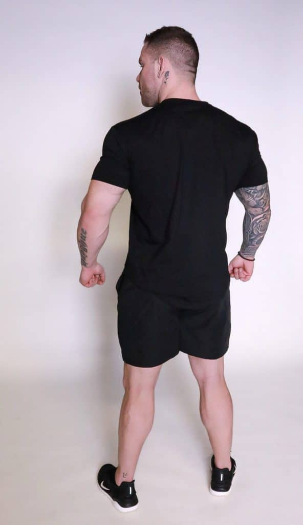 A muscular man with tattoos, wearing the Genesis Tee V2, black shorts, and black sneakers, stands facing away from the camera against a plain white background.