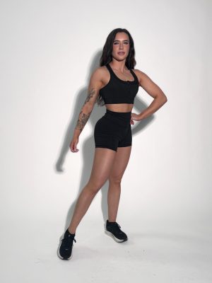 A woman with long dark hair, wearing a black sports bra, black shorts, and black sneakers, stands confidently against a plain white background. She has a tattoo on her left arm and poses with one hand on her hip.