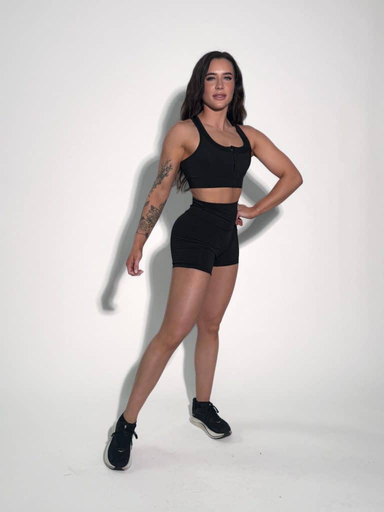A woman with long dark hair, wearing a black sports bra, black shorts, and black sneakers, stands confidently against a plain white background. She has a tattoo on her left arm and poses with one hand on her hip.