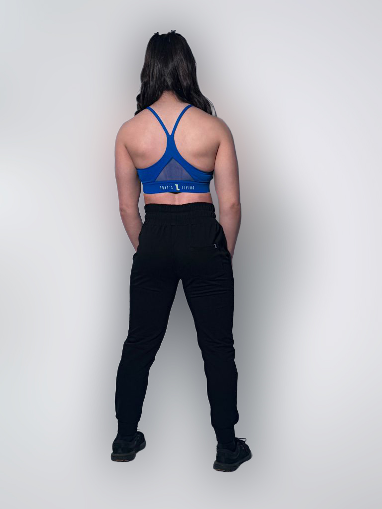 A person with long dark hair stands facing away, wearing a bright blue sports bra, black jogger pants, and black sneakers against a plain light background.
