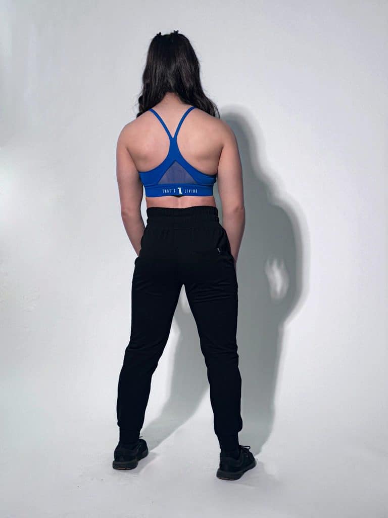 A person with long dark hair stands facing away, wearing a bright blue sports bra, black jogger pants, and black sneakers against a plain light background.