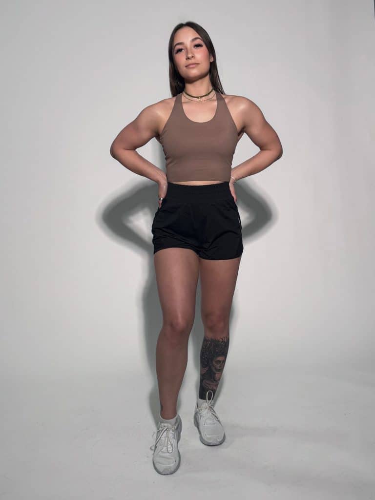 A young woman stands confidently with hands on hips, wearing a brown crop top, black shorts, and white sneakers. She has long brown hair and a tattoo on her left leg. The background is plain and light-colored.