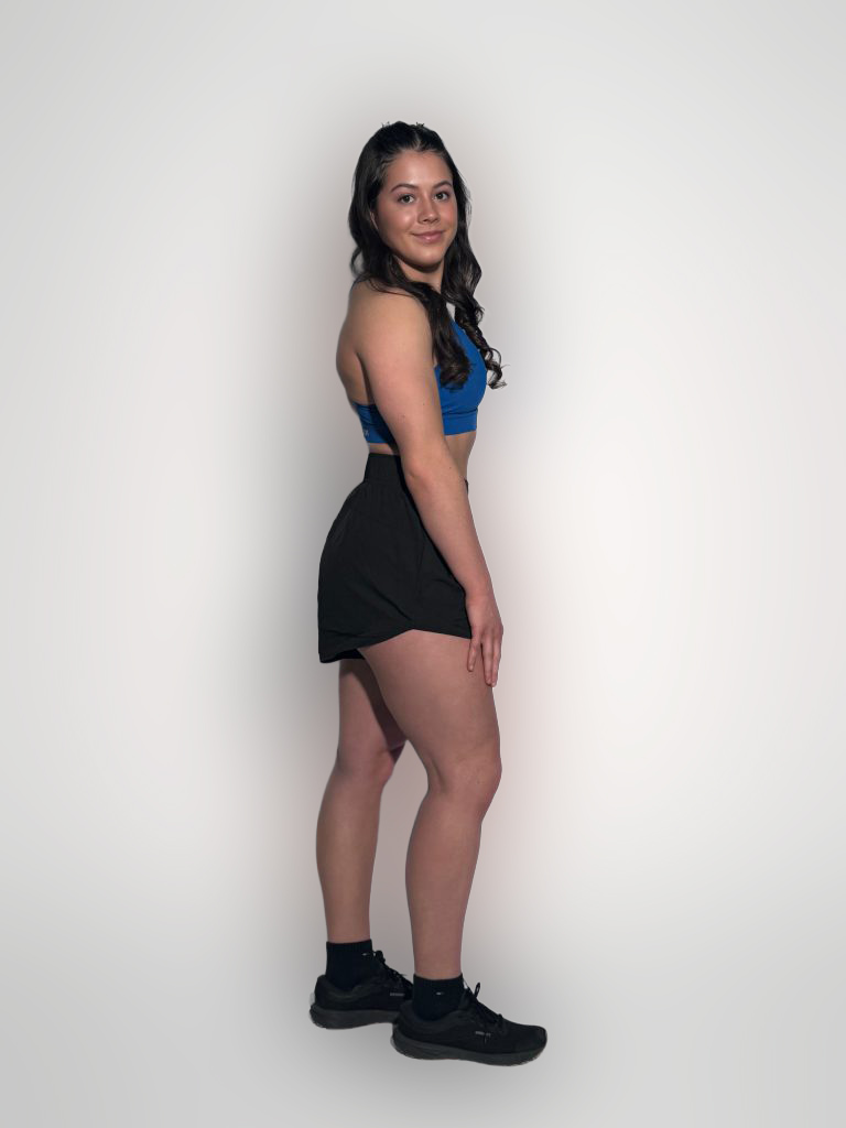 A woman with long dark hair stands sideways against a plain white background, wearing GLYDE Shorts with a blue sports bra, black socks, and black sneakers. She looks at the camera and smiles slightly.