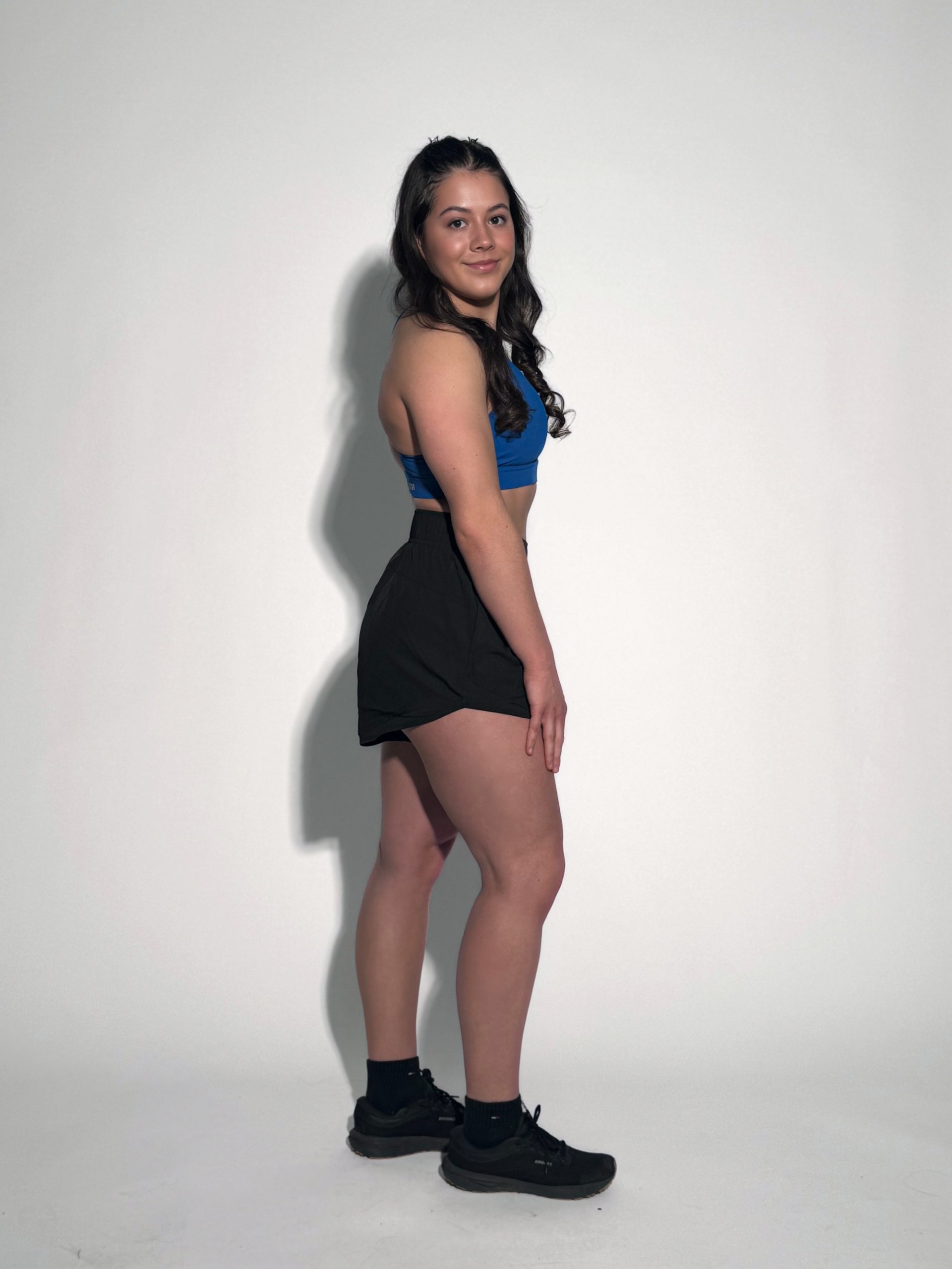 A woman with long dark hair stands sideways against a plain white background, wearing GLYDE Shorts with a blue sports bra, black socks, and black sneakers. She looks at the camera and smiles slightly.