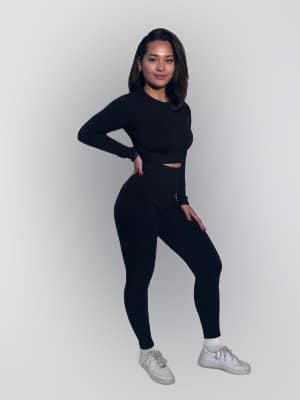 A woman stands against a plain white background, wearing a black long-sleeve top, matching black leggings, white socks, and white sneakers. She poses with one hand on her hip and smiles slightly at the camera.