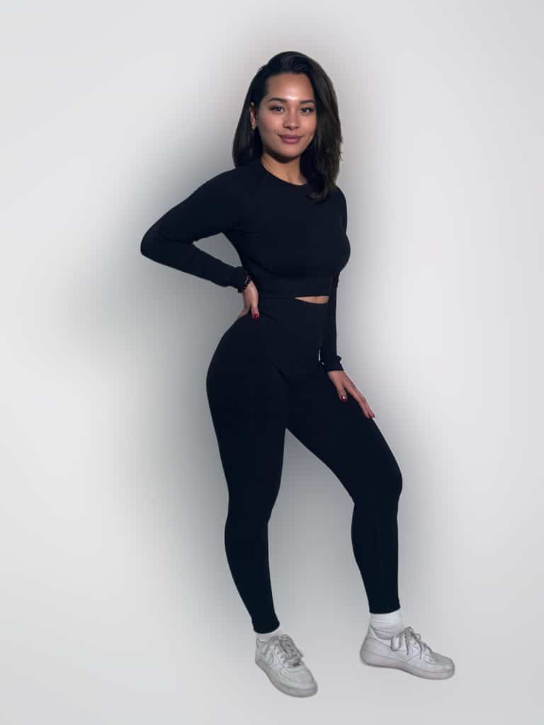 A woman stands against a plain white background, wearing a black long-sleeve top, matching black leggings, white socks, and white sneakers. She poses with one hand on her hip and smiles slightly at the camera.