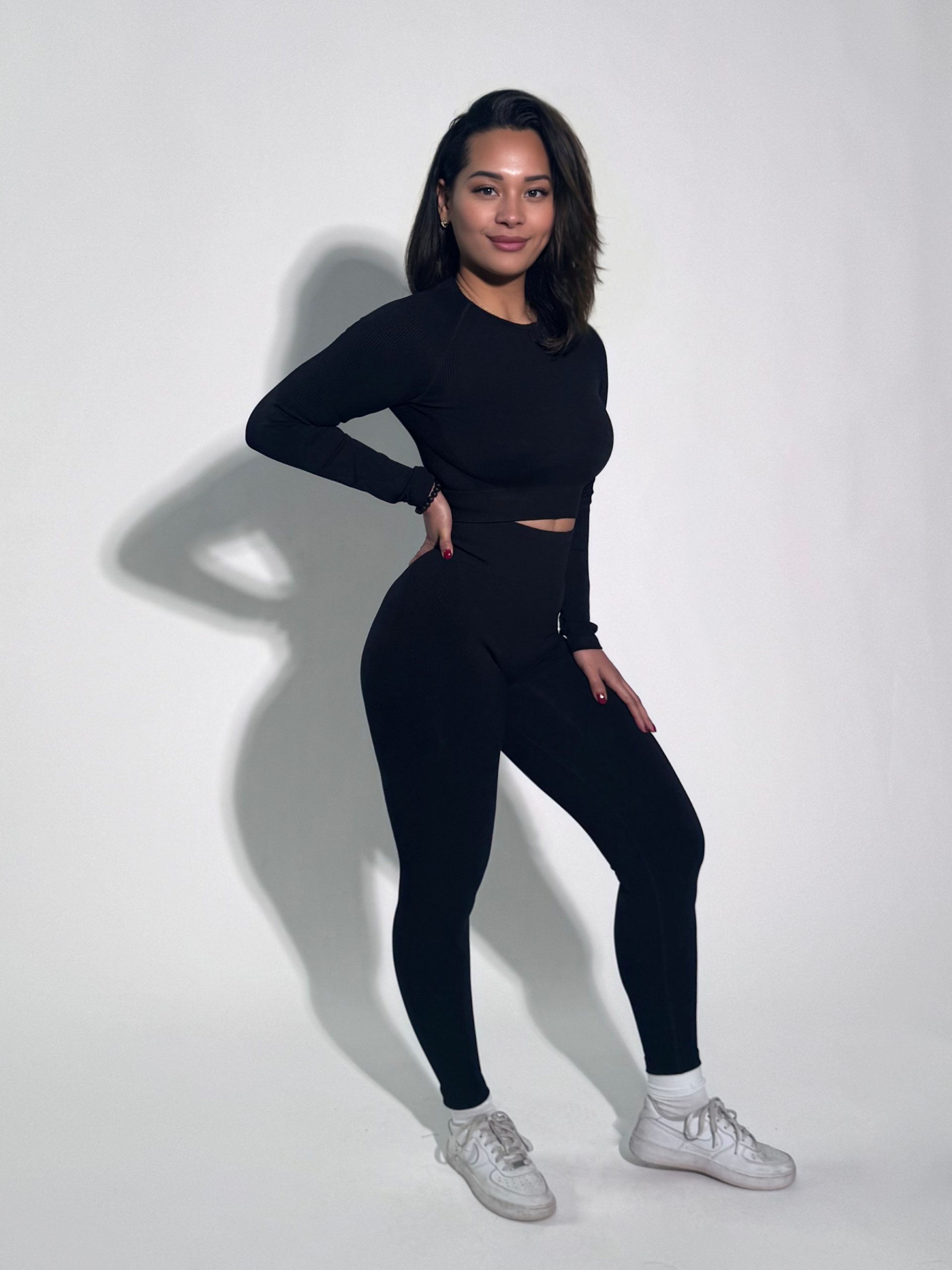 A woman stands against a plain white background, wearing a black long-sleeve top, matching black leggings, white socks, and white sneakers. She poses with one hand on her hip and smiles slightly at the camera.