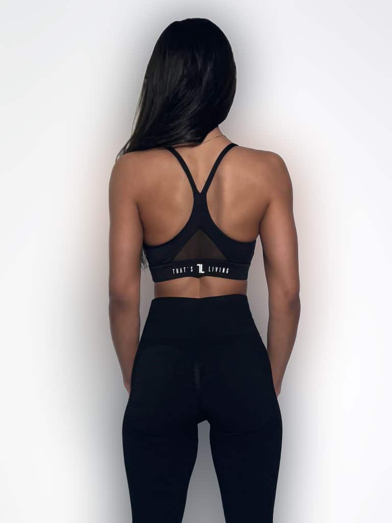 A woman with long dark hair stands facing away, wearing a black sports bra and black leggings against a plain white background. The sports bra has a racerback design and the text "THAT'S L LIVING" on the band.
