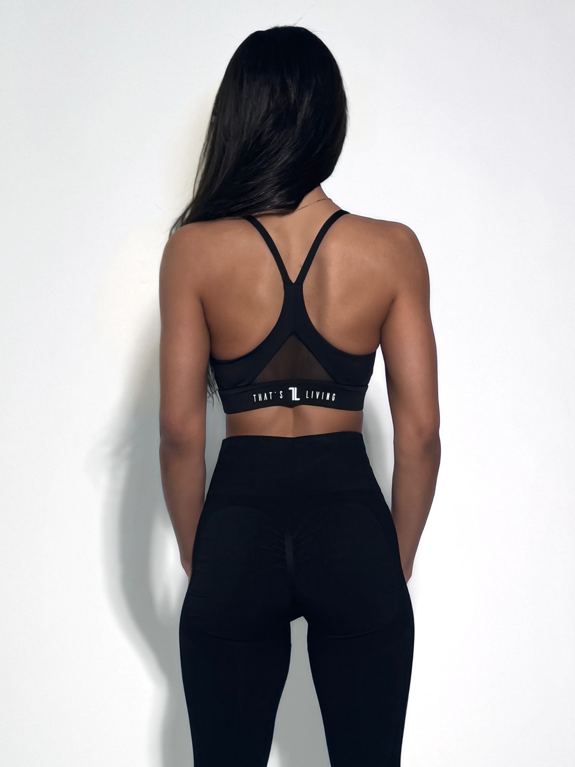 A woman with long dark hair stands facing away, wearing a black sports bra and black leggings against a plain white background. The sports bra has a racerback design and the text "THAT'S L LIVING" on the band.