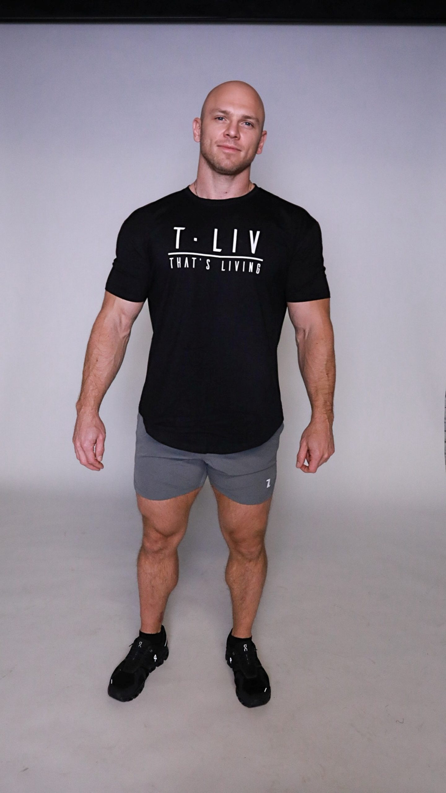 A muscular, bald man in a black Genesis Tee, gray shorts, and black sneakers stands against a plain light gray background, facing the camera with a neutral expression.