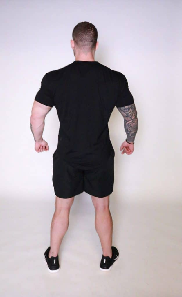 A muscular man with a tattooed right arm stands against a white background, facing away from the camera, wearing a black Genesis Tee V2, black shorts, and black sneakers.