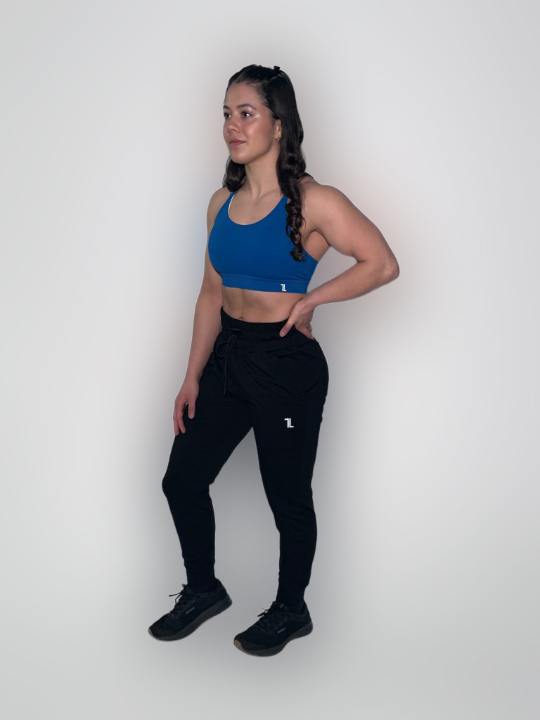 A woman in a blue sports bra and black joggers stands against a plain white background, with her left hand on her hip and her right arm relaxed by her side. She is wearing black sneakers and has long, dark hair in a braid.