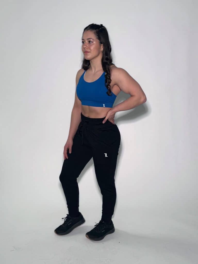 A woman in a blue sports bra and black joggers stands against a plain white background, with her left hand on her hip and her right arm relaxed by her side. She is wearing black sneakers and has long, dark hair in a braid.