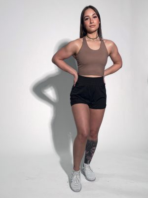 A woman poses confidently in a sleeveless brown top, black shorts, and white sneakers. She has a tattoo on her left leg and is standing against a plain light background with her hands on her hips.