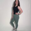 A woman with curly brown hair smiles against a plain light background, wearing a LuxeForm Tank, green leggings, and white sneakers, with one hand on her hip.