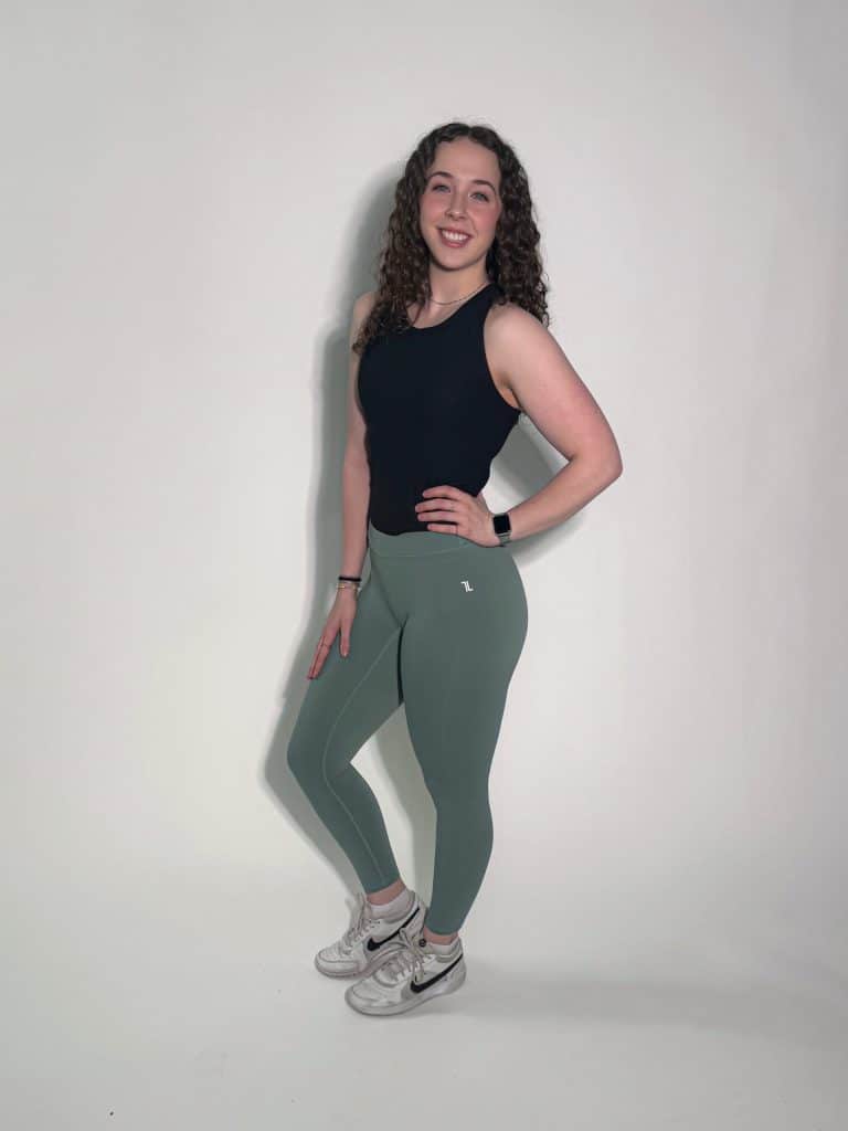A woman with curly brown hair smiles against a plain light background, wearing a LuxeForm Tank, green leggings, and white sneakers, with one hand on her hip.