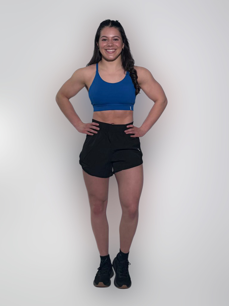 A woman with long dark hair in a braid smiles with her hands on her hips. She wears black GLYDE Shorts, a blue sports bra, and black athletic shoes, standing against a plain white background.