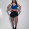 A woman with long dark hair in a braid smiles with her hands on her hips. She wears black GLYDE Shorts, a blue sports bra, and black athletic shoes, standing against a plain white background.