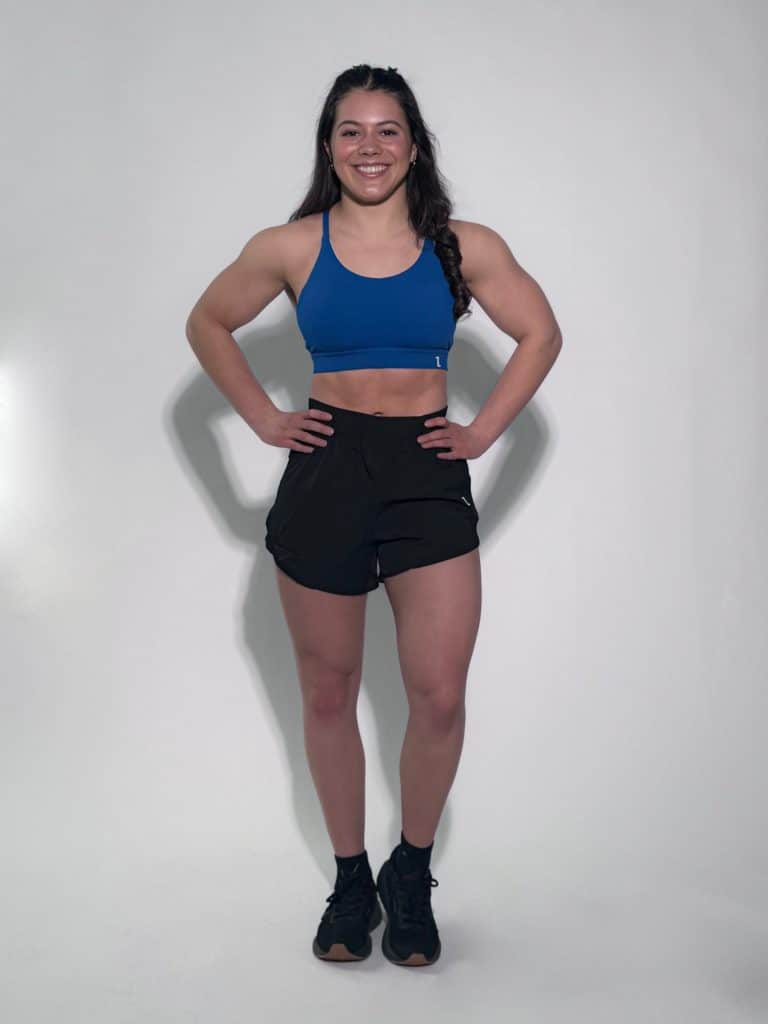 A woman with long dark hair in a braid smiles with her hands on her hips. She wears black GLYDE Shorts, a blue sports bra, and black athletic shoes, standing against a plain white background.