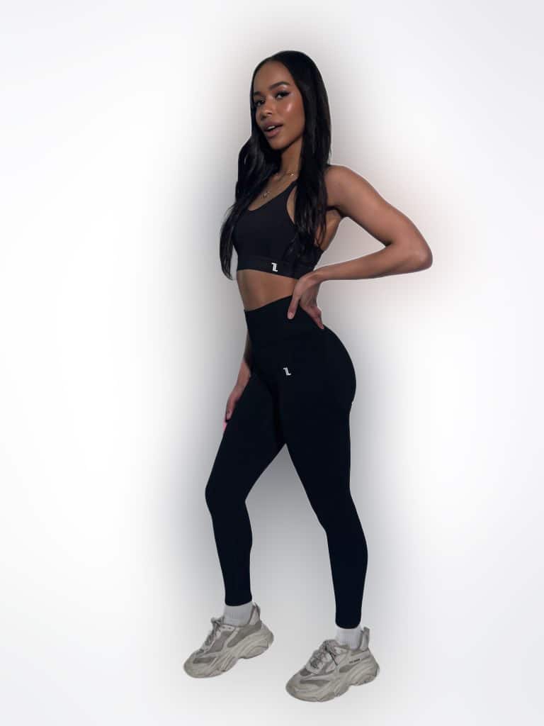 A woman poses against a plain white background, wearing a black sports bra, black leggings, and chunky white sneakers. She has long dark hair and her hand rests on her hip.