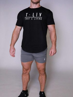 A fit, bald man stands against a plain background, wearing a black Genesis Tee with "T. LIV THAT'S LIVING" text, gray shorts, and black athletic shoes. He looks confident and relaxed.