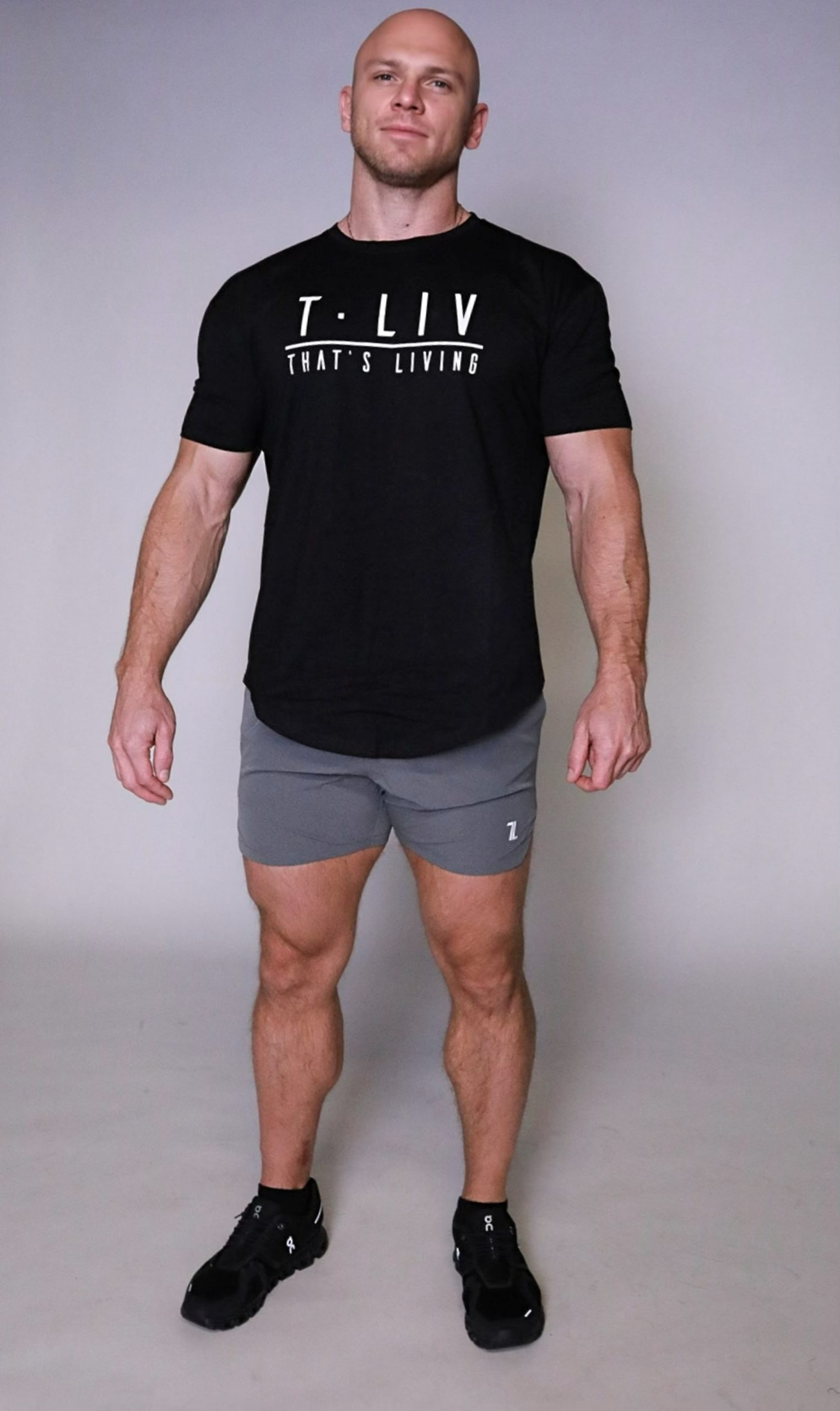 A fit, bald man stands against a plain background, wearing a black Genesis Tee with "T. LIV THAT'S LIVING" text, gray shorts, and black athletic shoes. He looks confident and relaxed.