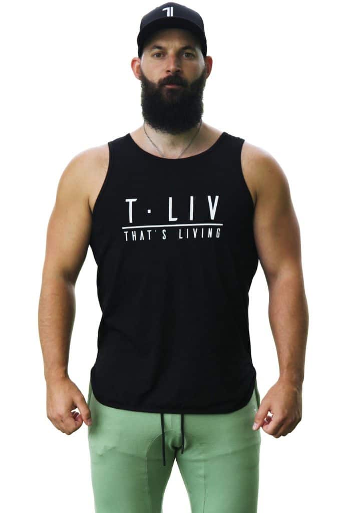 A muscular man with a full beard wears a black cap, a black tank top that says "T-LIV THAT'S LIVING," and light green pants. He stands facing forward against a white background.