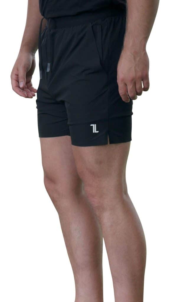 A person wearing black athletic shorts with a small white logo on the left leg stands against a white background; only the lower torso and legs are visible.