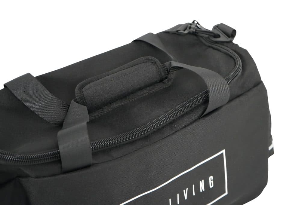 Close-up of the TRIARC Duffle Bag in black with a padded handle, zipper, and black straps. White text is partially visible on the side. The background is plain white.