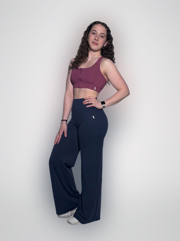A woman with long curly hair wears a mauve sports bra, high-waisted navy blue wide-leg pants, and white sneakers, standing against a plain light background and posing with one hand on her hip.