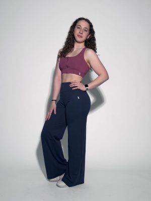 A woman with long curly hair wears a mauve sports bra, high-waisted navy blue wide-leg pants, and white sneakers, standing against a plain light background and posing with one hand on her hip.