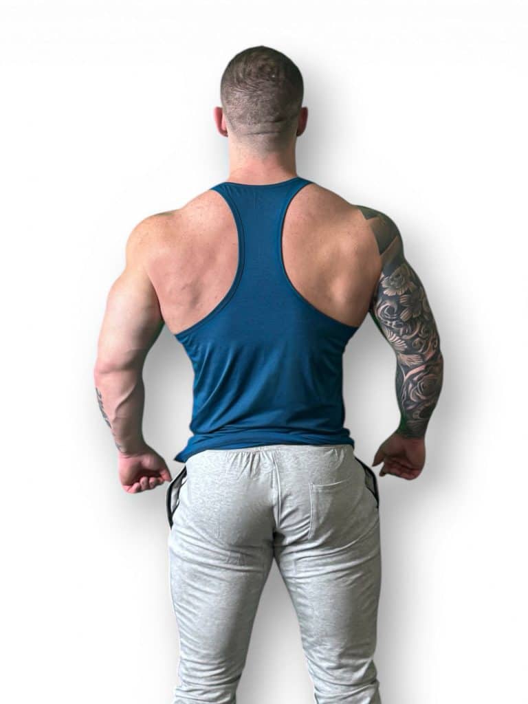 A muscular man with a tattooed right arm is shown from behind, wearing a blue tank top and light gray pants, standing against a white background.