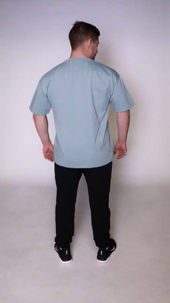A man stands facing away from the camera in a well-lit studio, wearing the light blue Equity Tee with tapered fit and scoop bottom, paired with black pants and black sneakers with white soles.