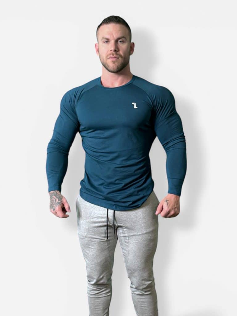 A muscular man wearing a fitted dark teal long-sleeve shirt and light gray jogger pants stands facing the camera against a plain white background.
